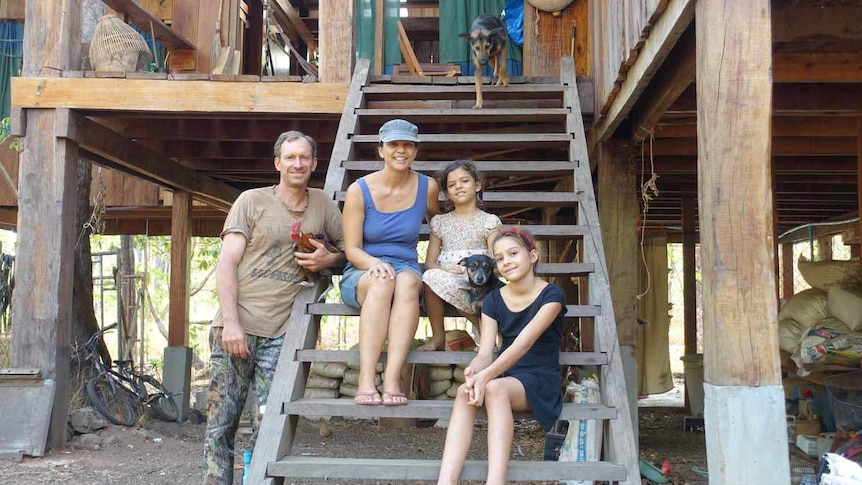 Ben and Sharyn Davis sit with their two daughers, a dog, and chicken on stairs outside their wooden home.