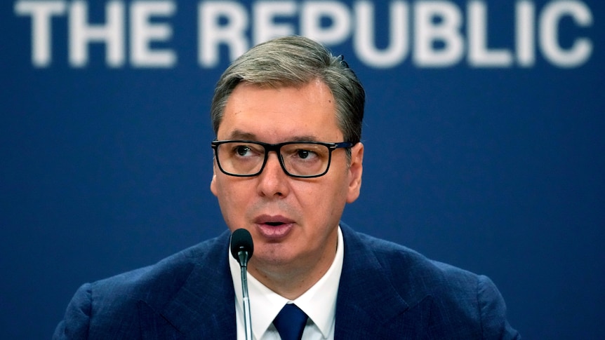 Close up of man in navy suit and glasses speaking into microphone.