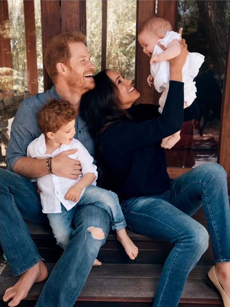 Meghan holds a giggling Lilibet in the air as Harry holds Archie on his knee