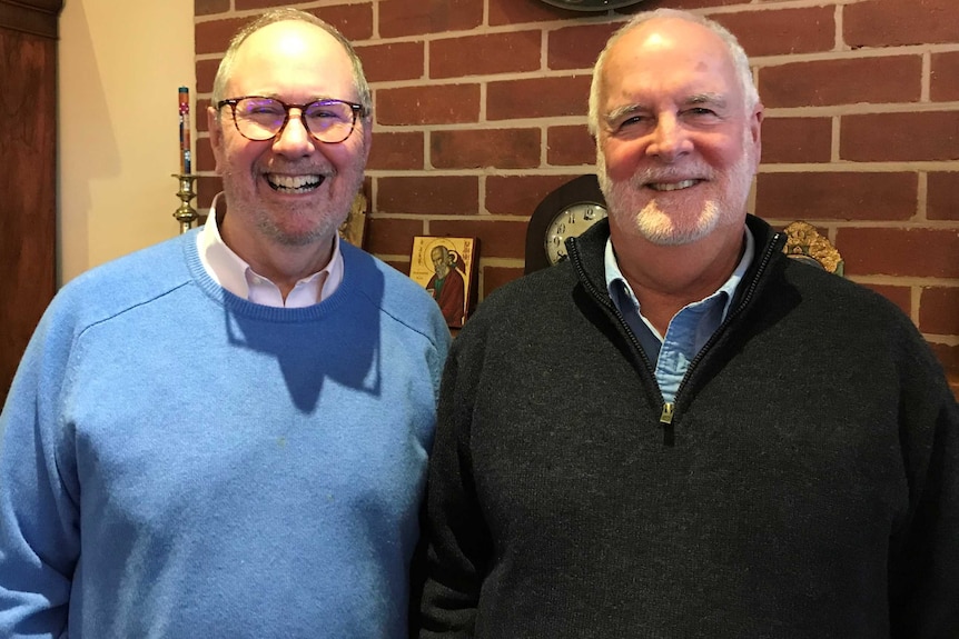 Two men smile at the camera, with Christian relics behind them on a mantle.