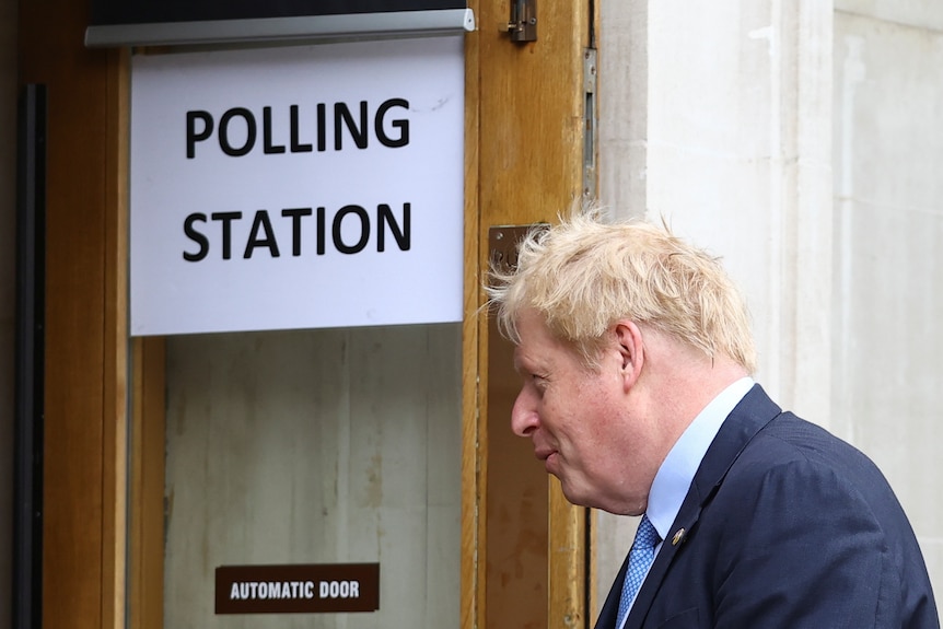 A man with blonde hair standing side on next to a sign that says Polling Station.