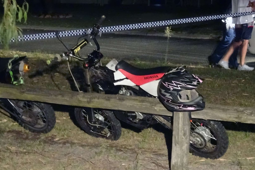 One of the motorcycles being ridden by the teenagers leaning against a wood barrier.