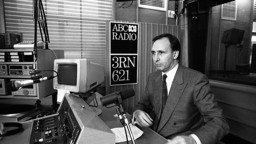 Man in suit sitting in front of a microphone in a radio studio with computers and panels on desk