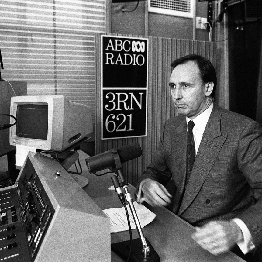 Man in suit sitting in front of a microphone in a radio studio with computers and panels on desk