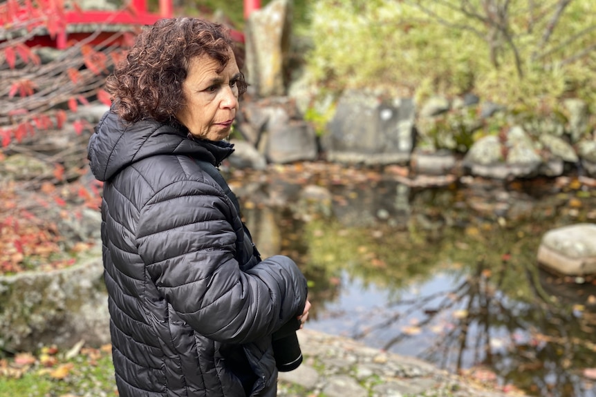 A woman in a puffer jacket stands in a garden with a camera around her neck