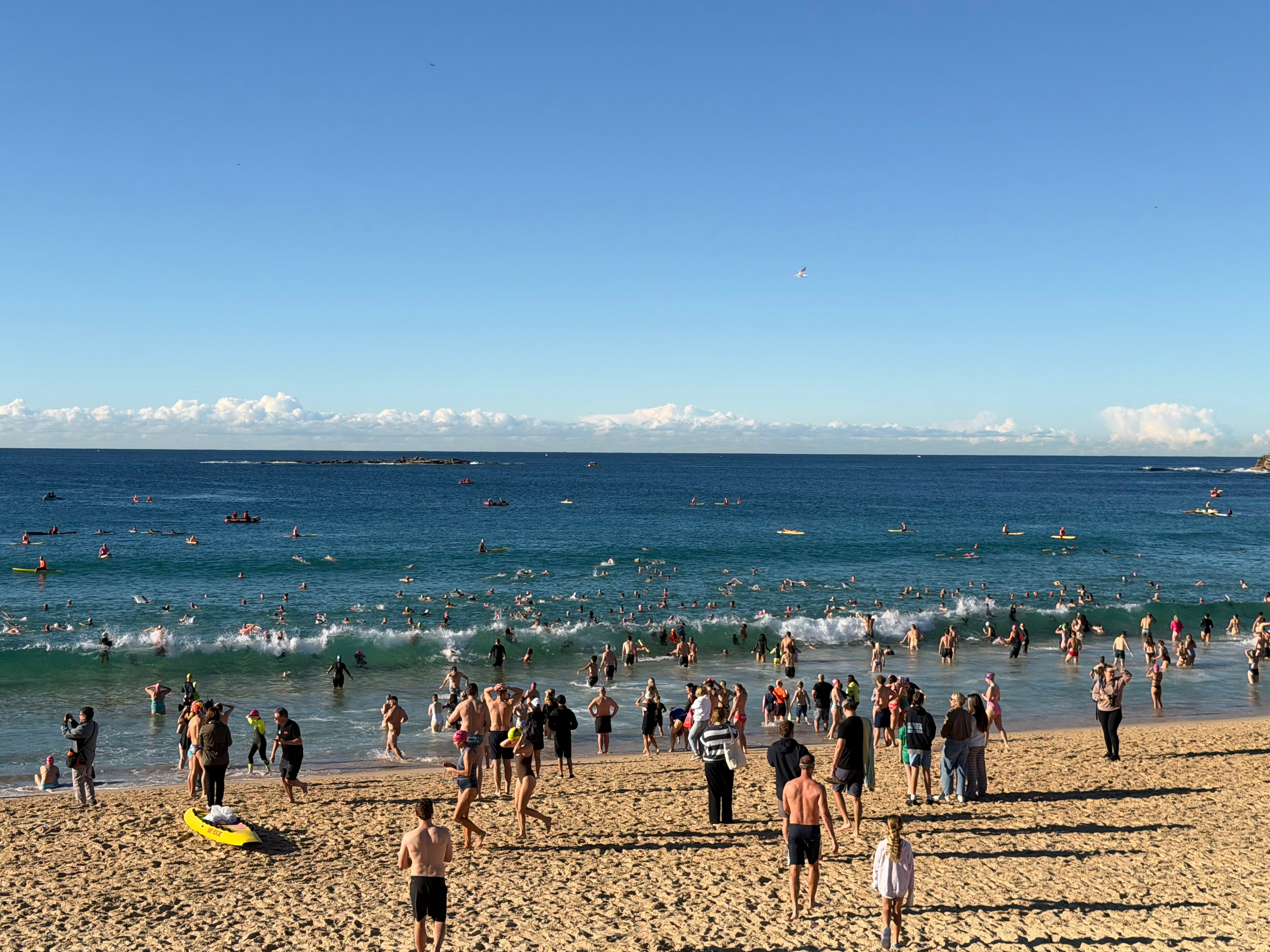 Swimmers stand watching on the sand as others enter the water on a bright morning.