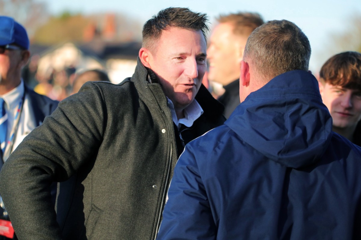 A man on a football pitch speaking to another man and smiling. They are surrounded by people celebrating.