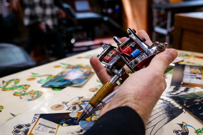 A hand holding an old tattoo machine, with old tattoo designs on a desk. 