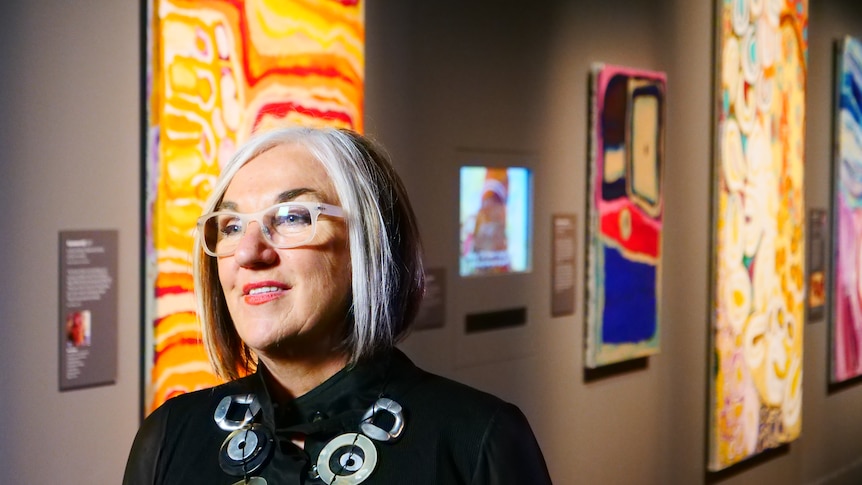 Margo Neale looks into the distance while standing in front of artwork from the Songlines exhibition.