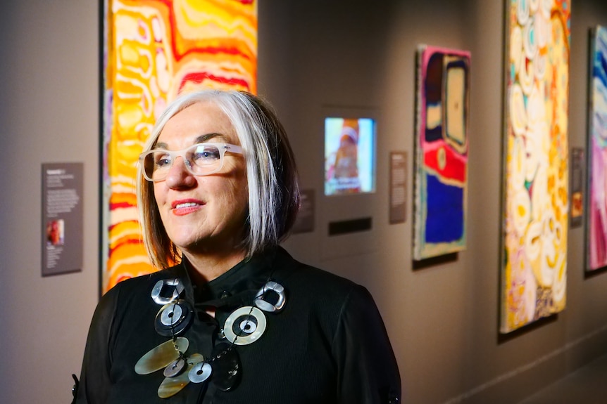 Margo Neale looks into the distance while standing in front of artwork from the Songlines exhibition.