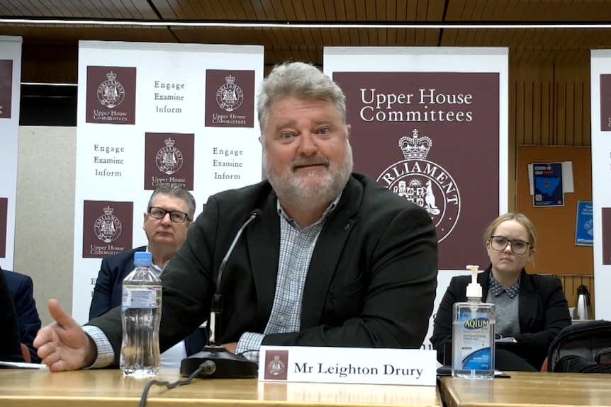 A man in a suit sits at a table, gesticulating as he speaks. New South Wales government logos appear in the background.