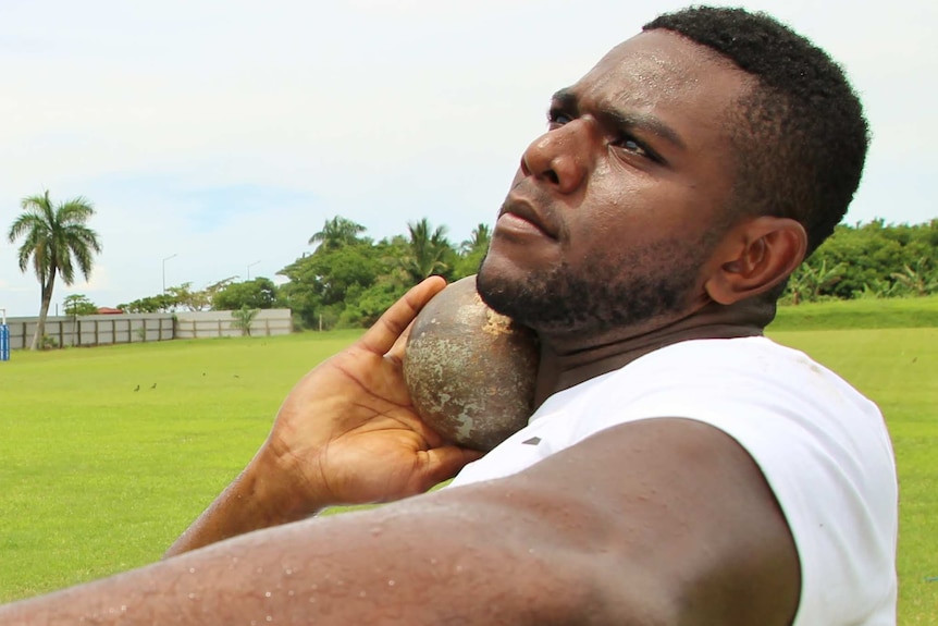 Athlete Mustafa William Fall holds a shot put under his chin, ready to throw.