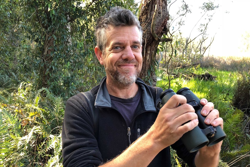 A middle-aged man with a greying beard stands in bushland and smiles as he holds a pair of binoculars.