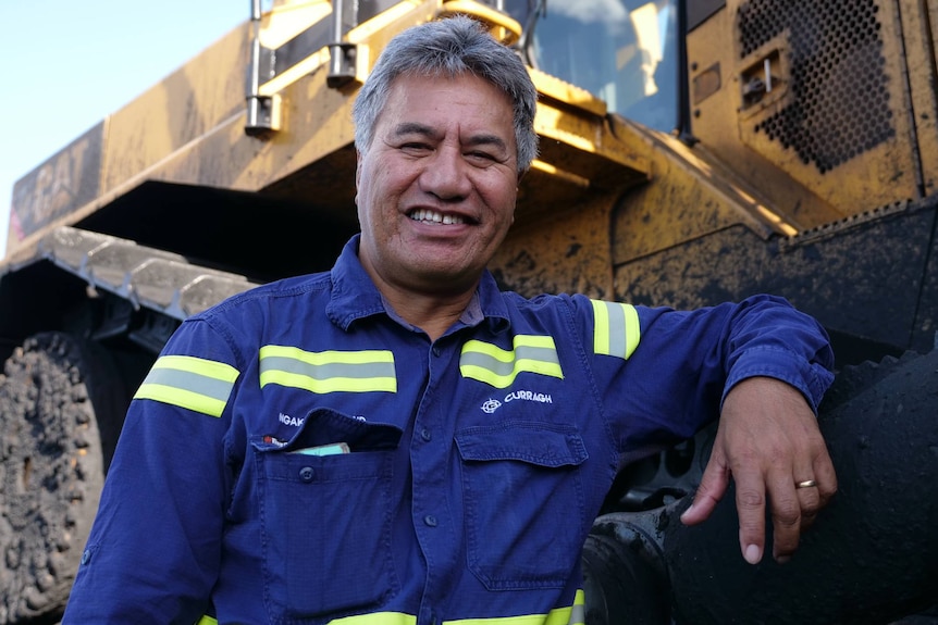 A man wearing hi-vis leans against a mining truck and smiles toward the camera