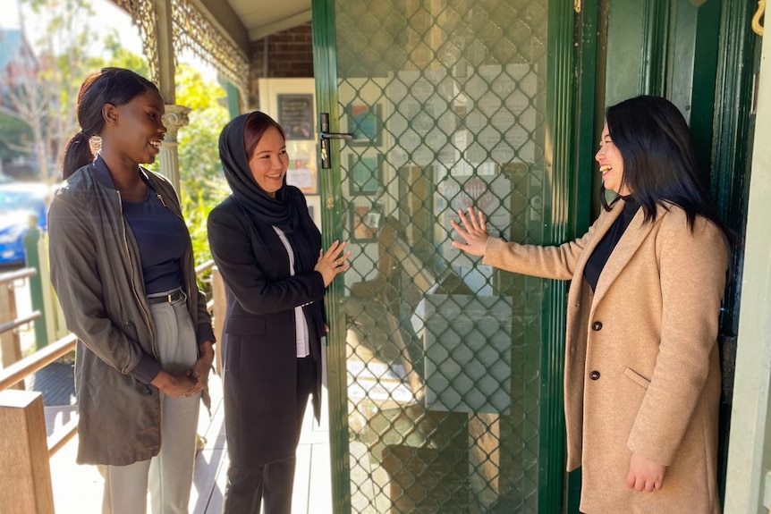 Three women stand at an open door smiling.