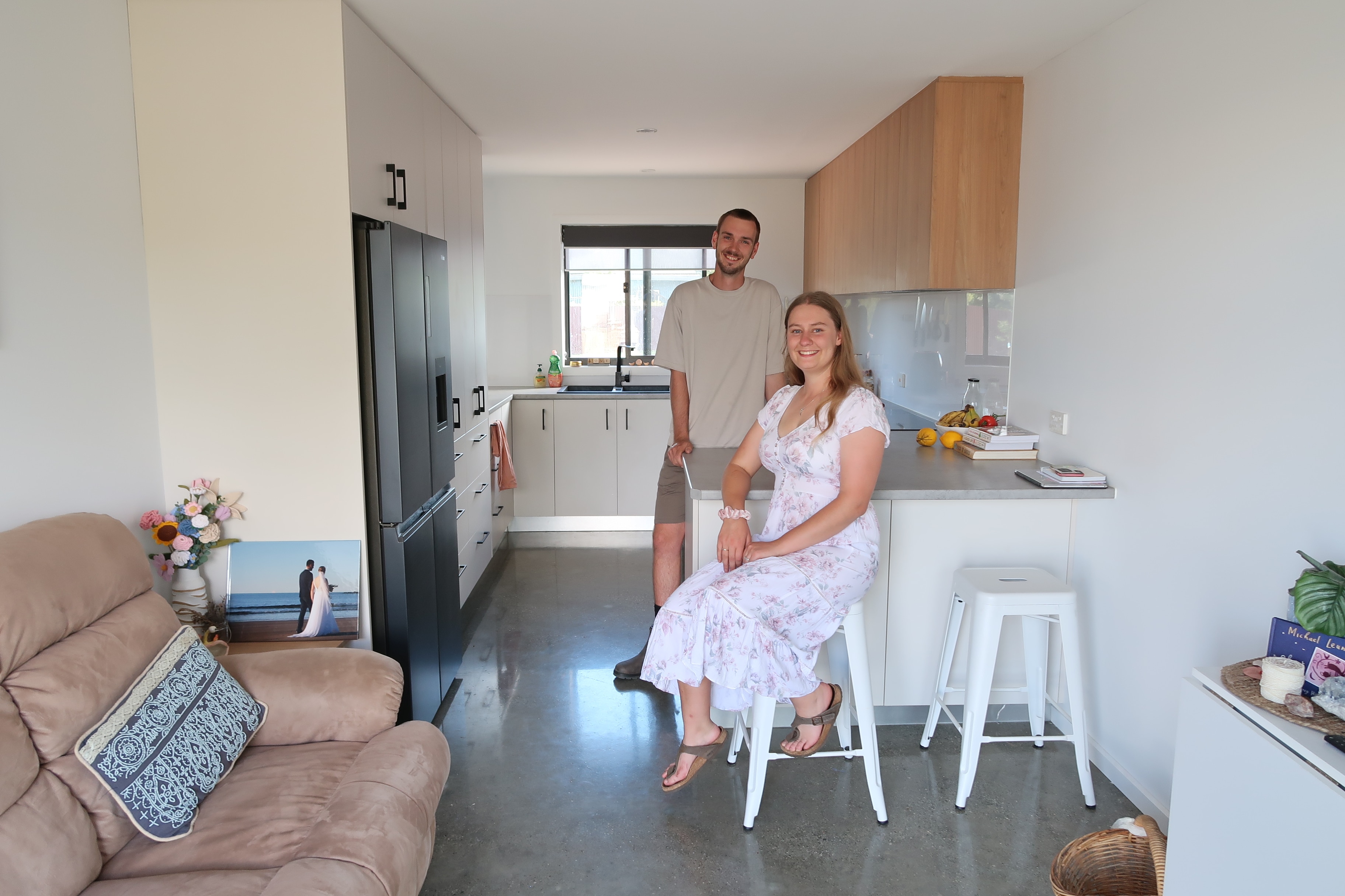 A young woman with long blond hair sits on a stool in a kitchen smiling. A man with short brown hair stands behind her.