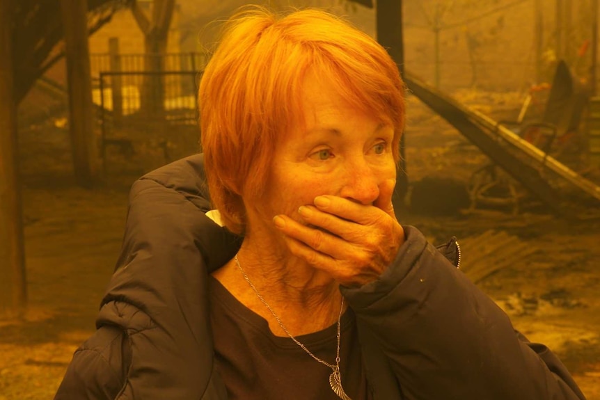 A woman puts her hand over her mouth and looks shocked surrounded by burned property and an orange glow
