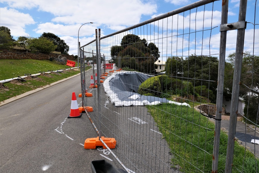 A construction fence blocking off an area of landslip-damaged land. The fence is one the road and a tarp covers the sunken area