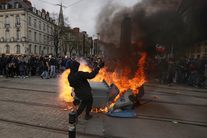 a man in black clothing stands next to a burning bin as protesters gather in Nantes