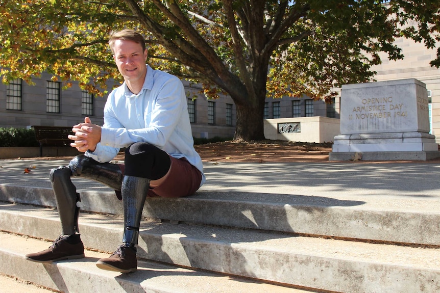 A man with two prosthetic legs sits of concrete steps, outside the leafy surrounds of the Australian War Memorial.