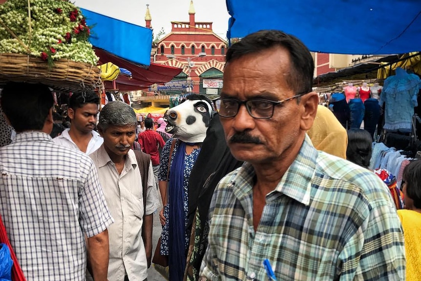 A woman walks through a busy market with a cow mask on.