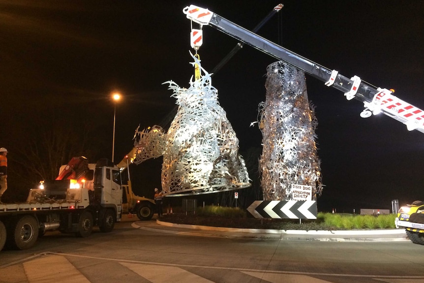 A crane lifts a metallic sculpture off a roundabout