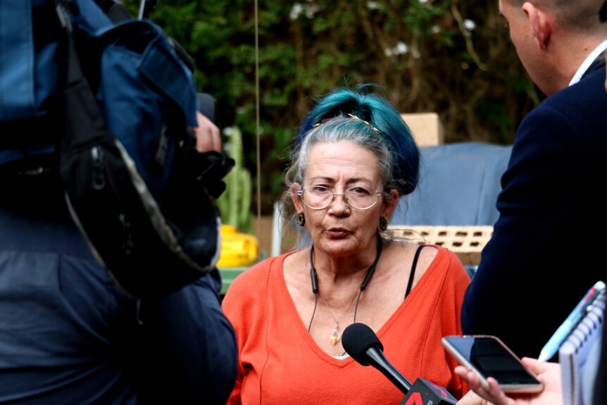 A woman with dyed blue hair wearing an orange top speaks to a group of reporters with trees in the background.