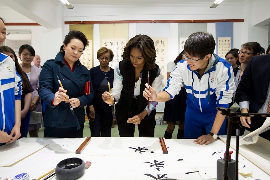 Peng Liyuan teaches Michelle Obama how to draw calligraphy, a male student standing next to Obama. 