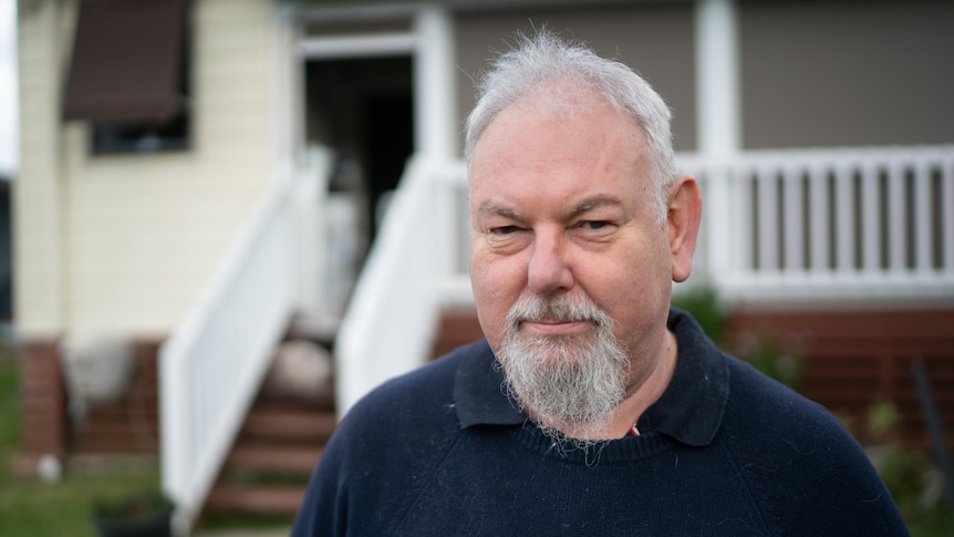 Man with grey beard standing out the front of his house looking at camera not smiling 