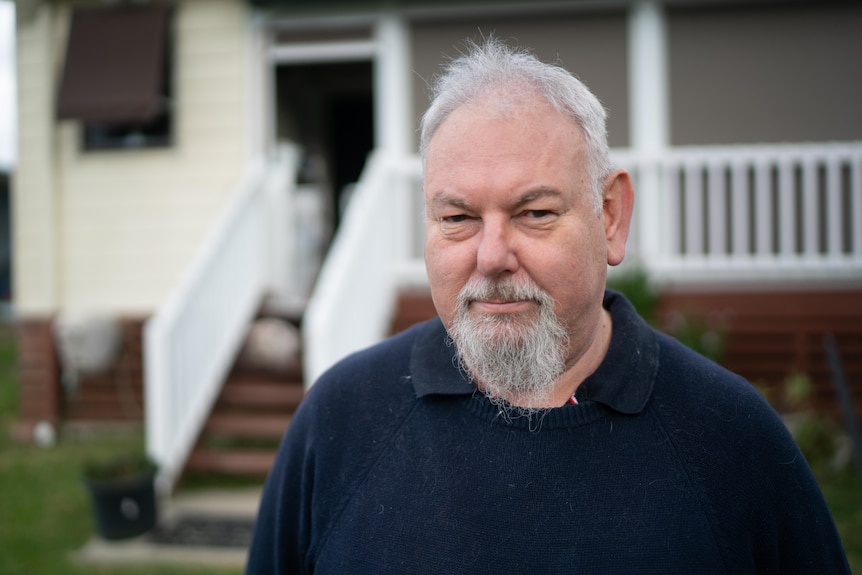 Man with grey beard standing out the front of his house looking at camera not smiling 