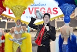 A man in an Elvis costume holds a rugby league ball like an American football, with women in showgirl costumes.