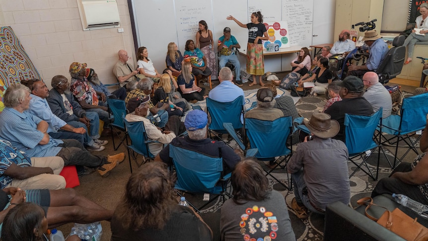 A group of people gathered in chairs around a whiteboard, with the coroner and several people at the front 