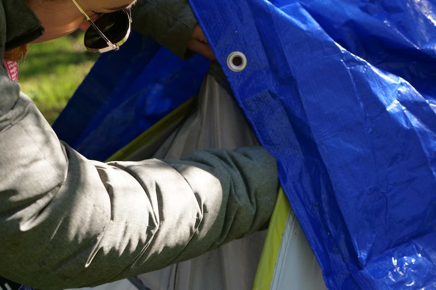 A woman's arm reaches inside a grey tent covered with a blue tarp