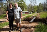 Andrew Gardyne and Else Seligmann hold hands while standing near a large hole in the ground with a plank of wood across it.