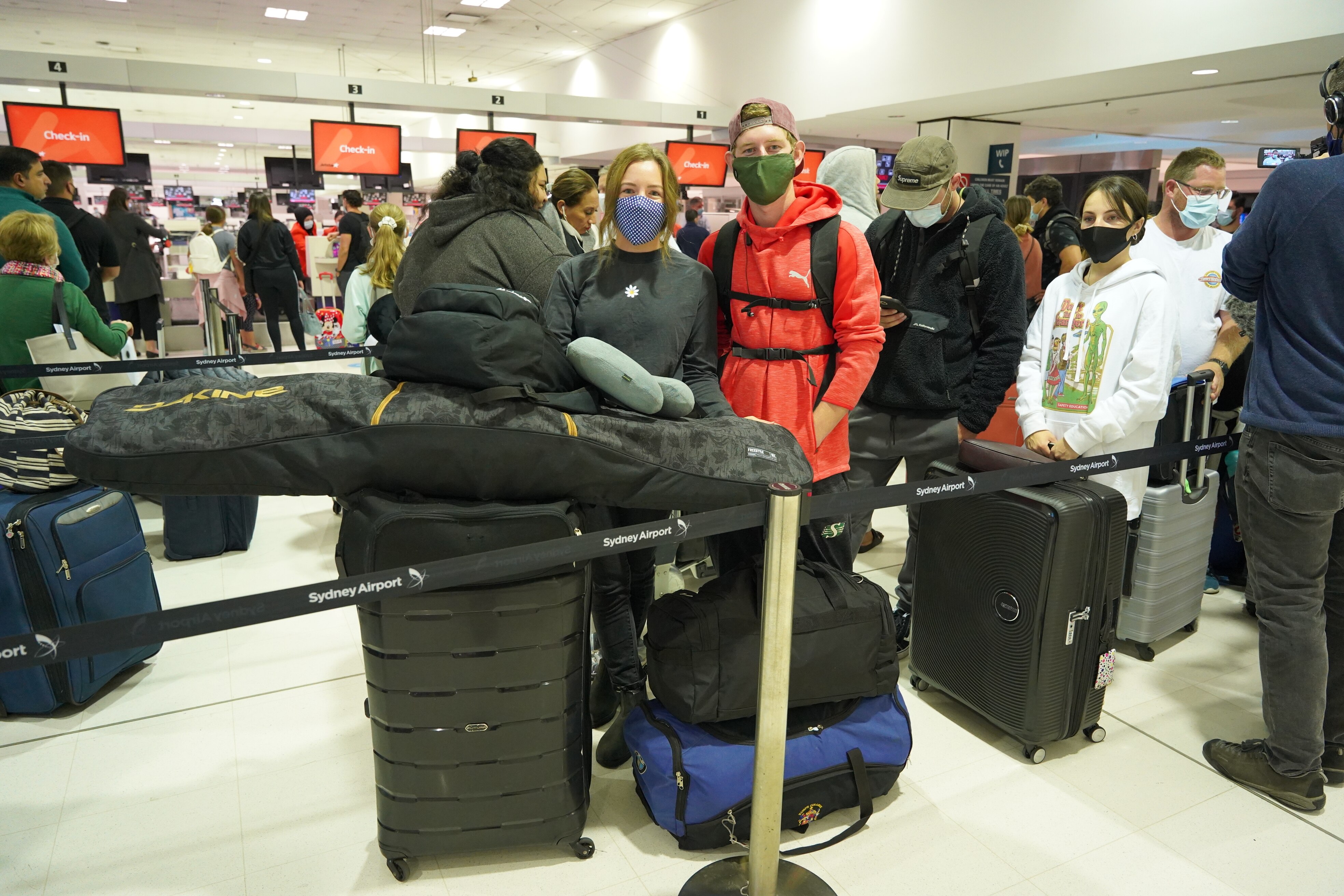 A woman and a man in a red jacket standing in the airport queue with several suitcases, both wearing a face mask.