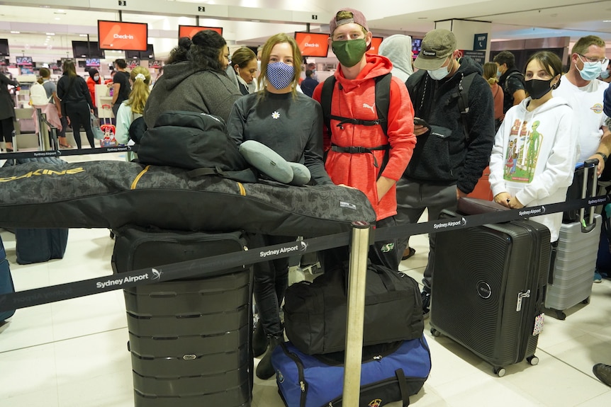 A woman and a man in a red jacket standing in the airport queue with several suitcases, both wearing a face mask.