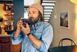 A wide portrait of Joel Edgerton in his home holding a camera and wearing beanie