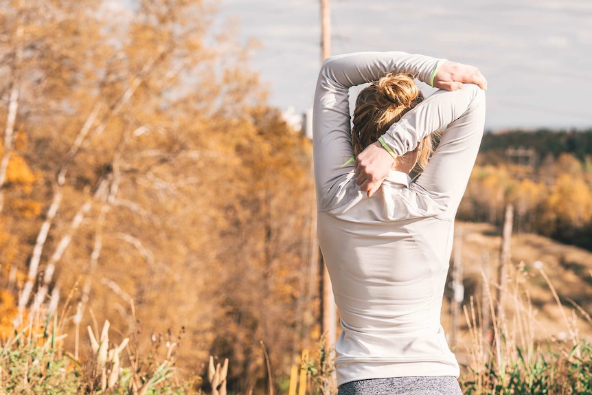 Woman in exercise clothes stretching her arms as she faces the bush to depict exercise as a way to help work stress.