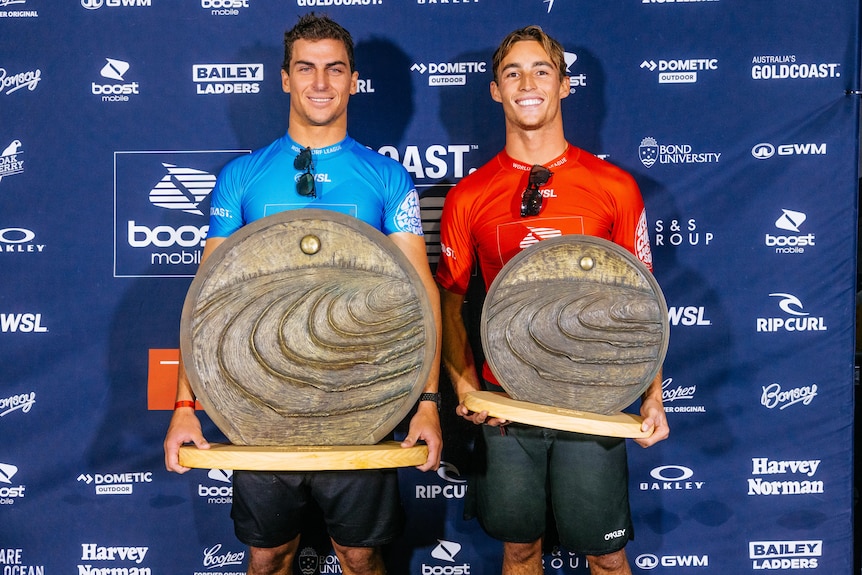 Two men in rash shirts stand next to each other, holding large trophies and smiling at the camera.