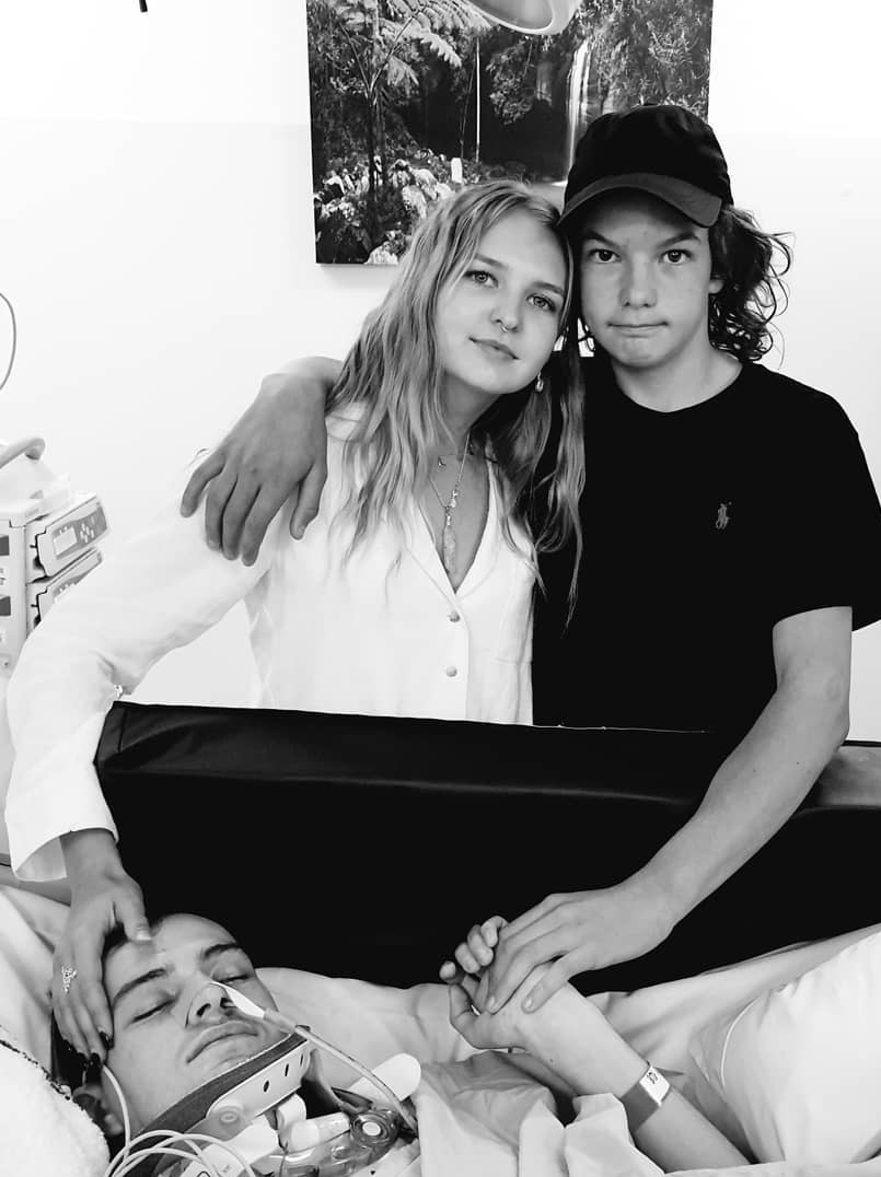 Young man sleeping in hospital connected to breathing apparatus with sister and brother hugging, holding his hand and head