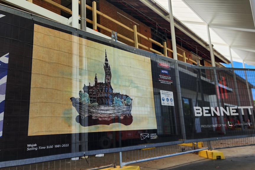 A large brick building with scaffolding and construction, surrounded by a temporary fence with an artwork on it.