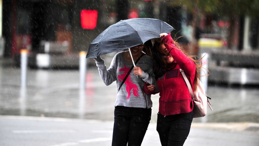 Two women cross the street in the rain with one protected under an umbrella and the other pulling it towards her.