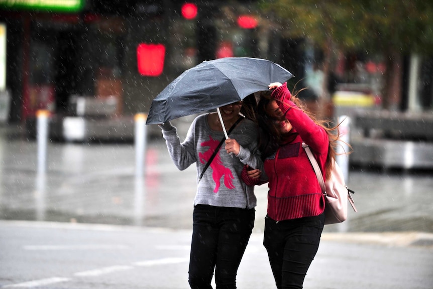 Two women cross the street in the rain with one protected under an umbrella and the other pulling it towards her.