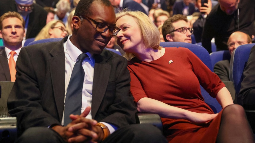 liz truss, in red, leans in to speak into kwarteng's ear in the audience of the annual conference