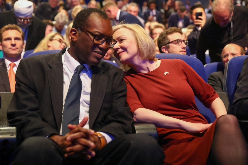 liz truss, in red, leans in to speak into kwarteng's ear in the audience of the annual conference