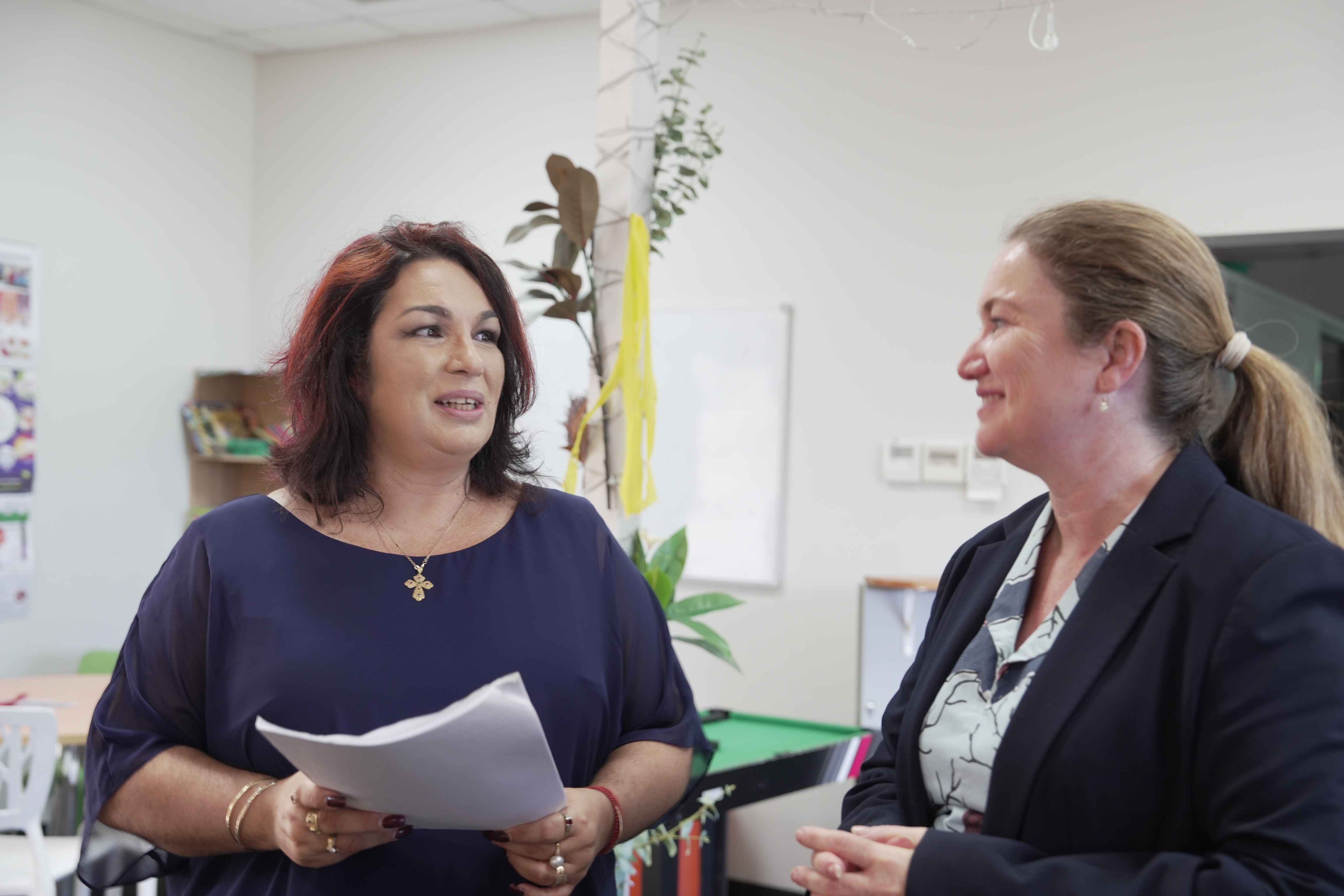 Duas mulheres conversando, uma delas com papéis nas mãos, em ambiente educacional.