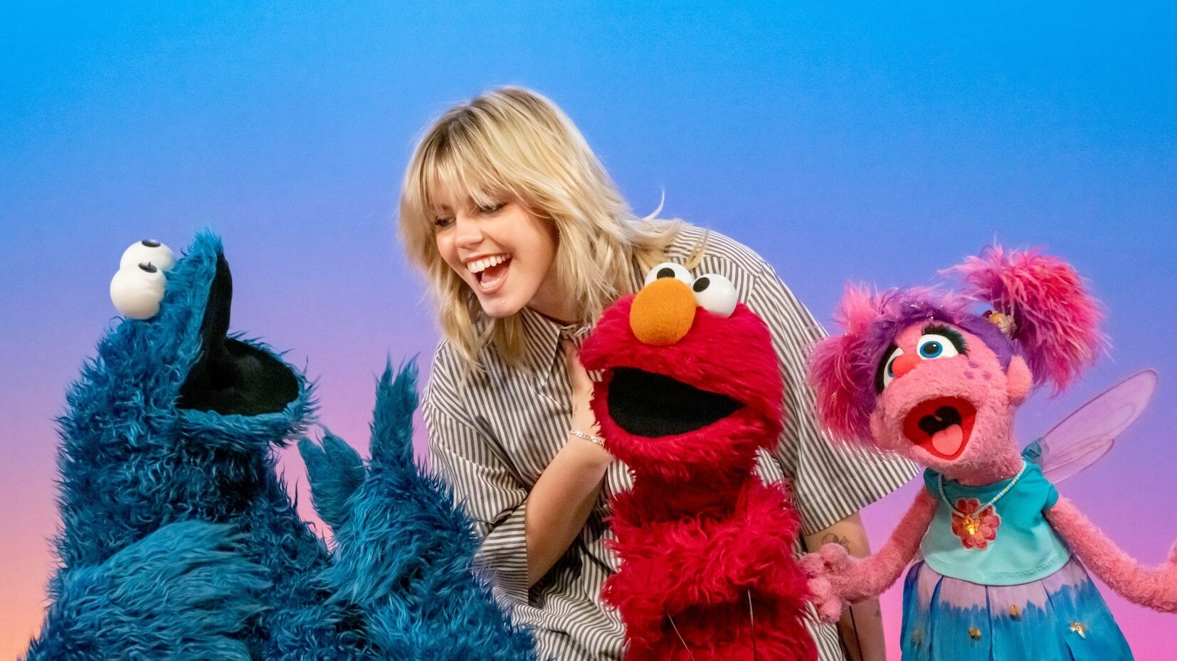 A blonde woman in a striped shirt leans down to talk to muppets - all are smiling