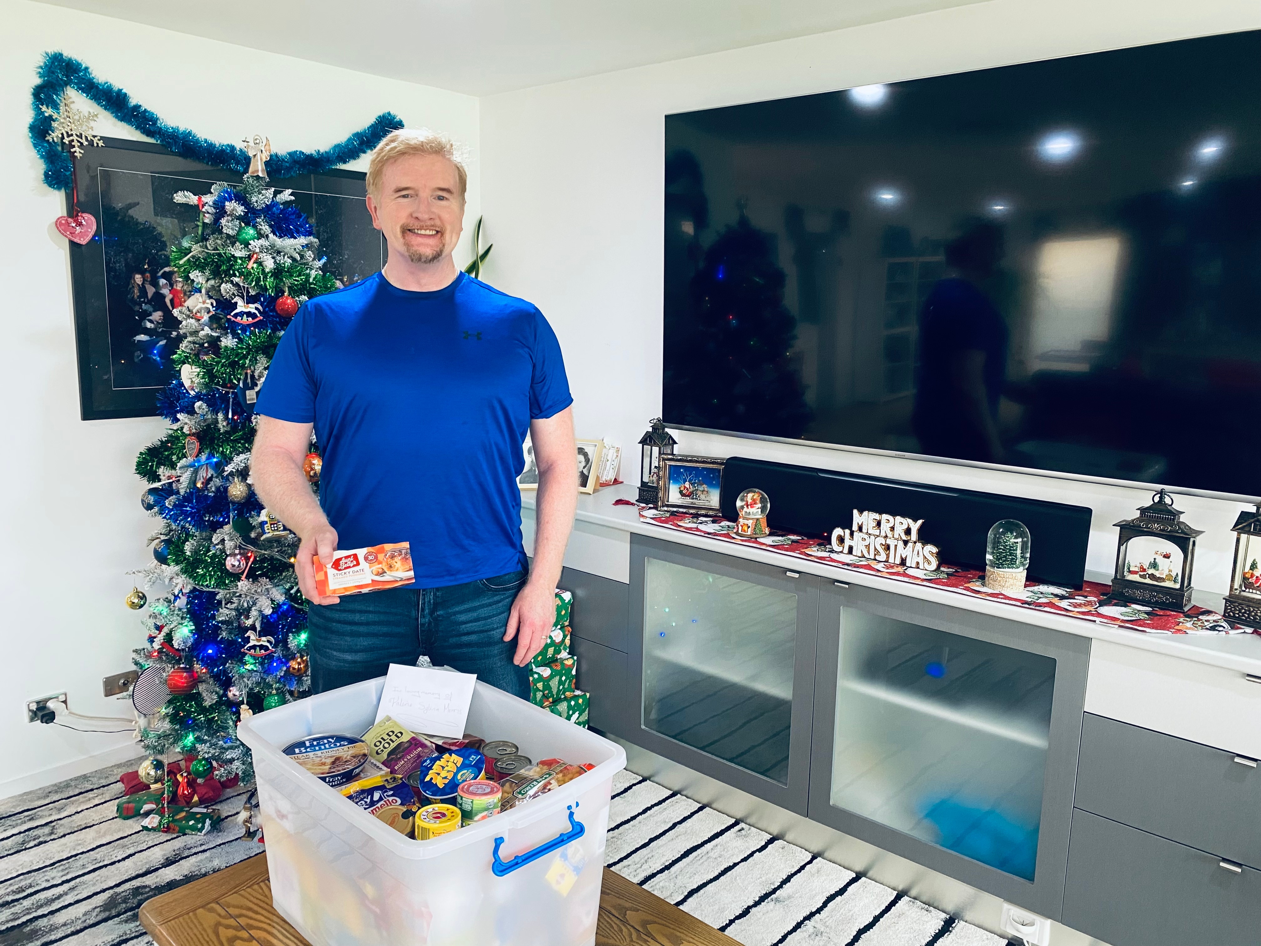A man in a blue t-shirt standing next to a Christmas tree and a tub of goods. 