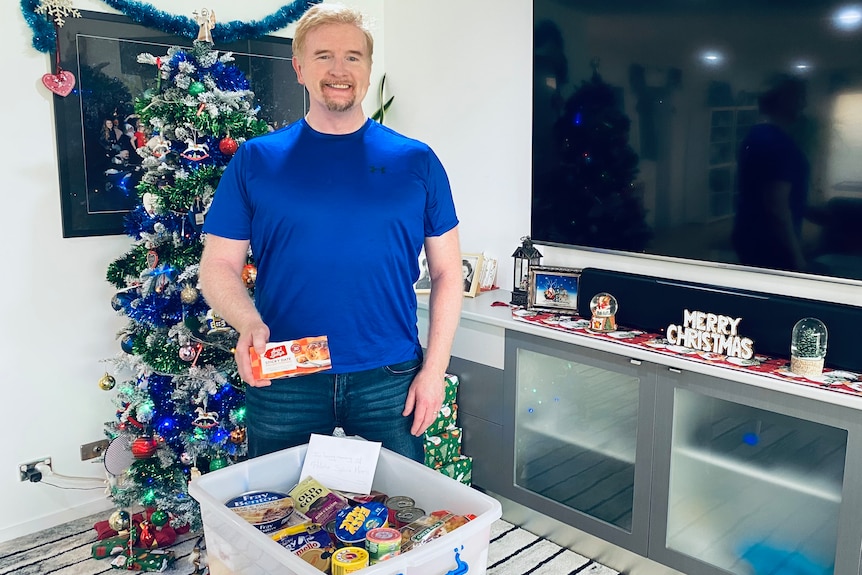 A man in a blue t-shirt standing next to a Christmas tree and a tub of goods. 