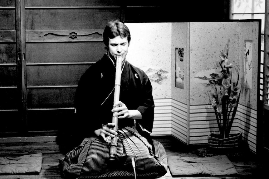 A man sits on a cushion on the floor playing the shakuhachi on Japanese TV.